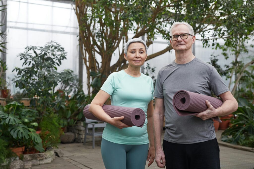 Corrective Exercise + Functional Strength Coaching Older couple with yoga mats standing in a serene greenhouse setting.